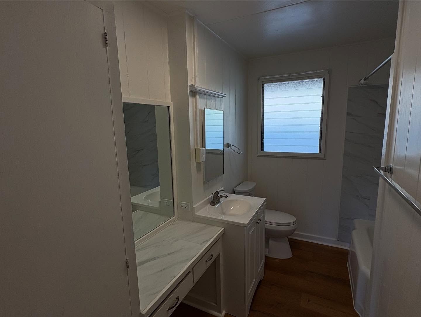 Bathroom with vanity, toilet, and bathtub. White walls and marble-patterned surfaces. Window with frosted glass.