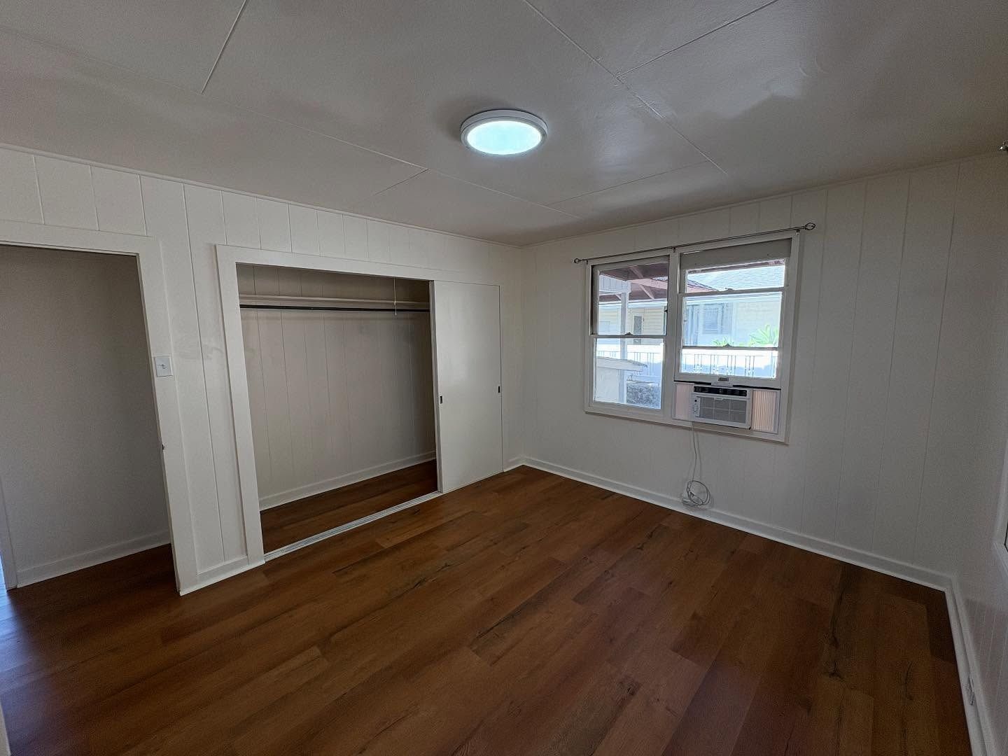 Empty room with closet and window, hardwood floors, white walls, and a circular ceiling light.