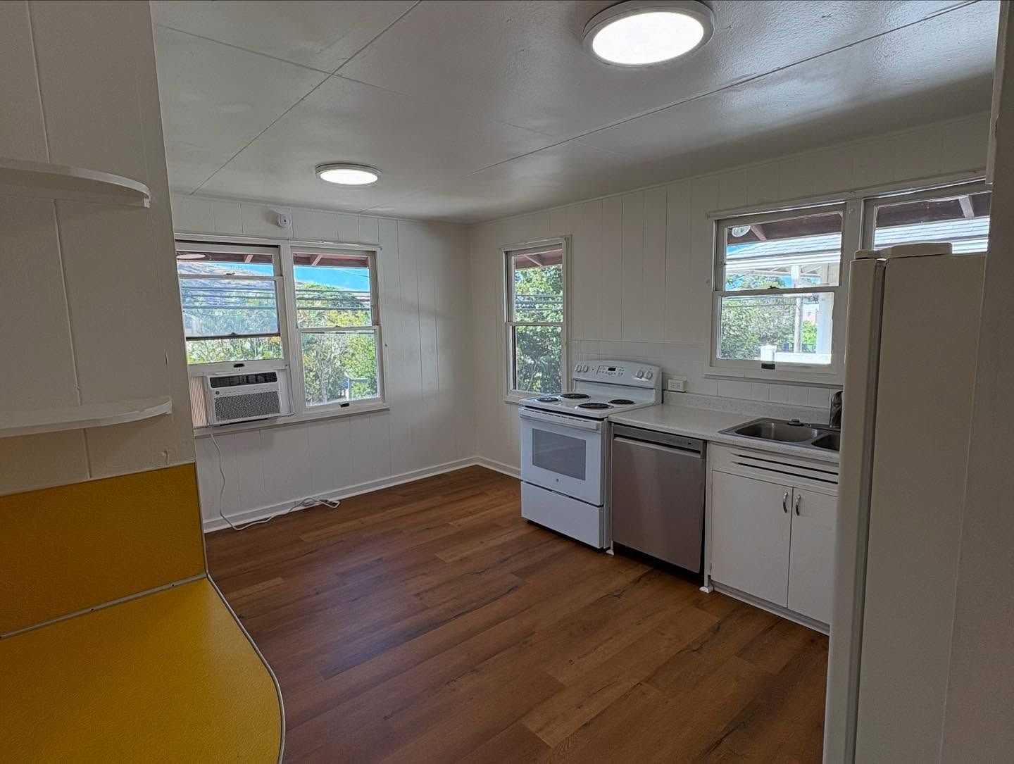 Kitchen with white cabinets, stainless steel appliances, and wood flooring. Three windows and overhead lighting.