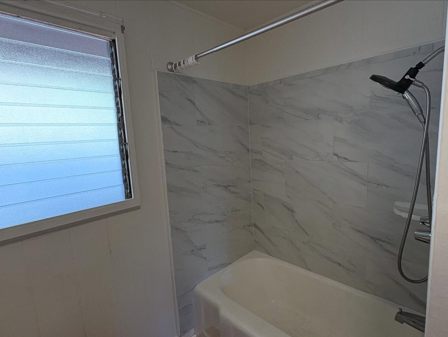 Bathroom with white bathtub, marble-look shower surround, showerhead, and window with frosted glass.