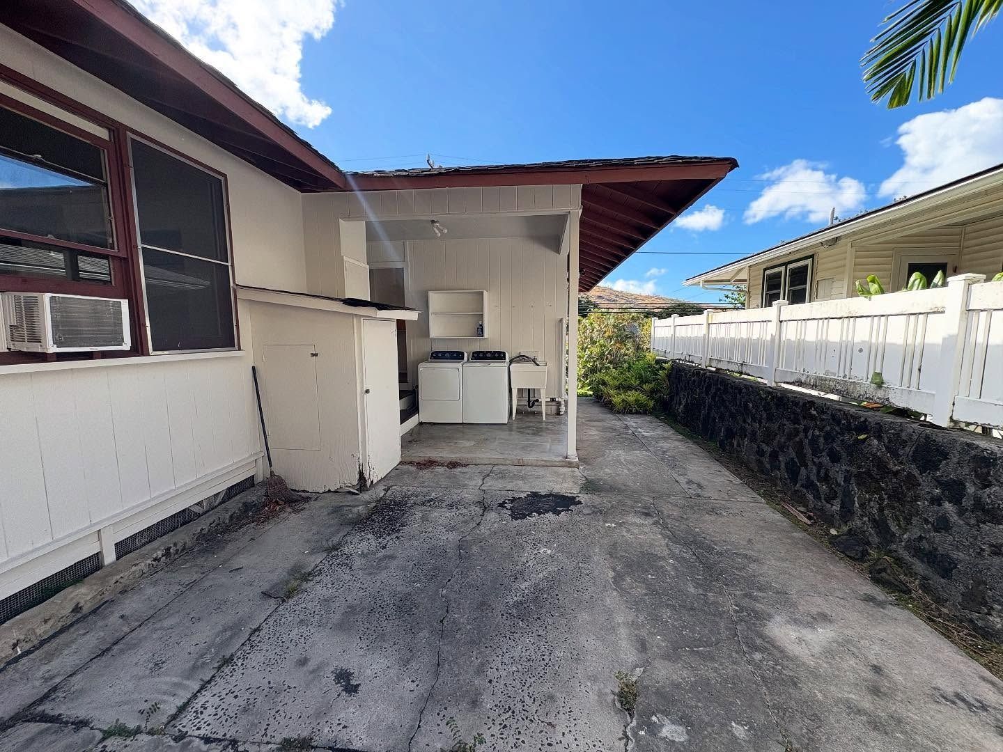 Driveway leading to a house with a carport and laundry area; white exterior, blue sky, and a fence.