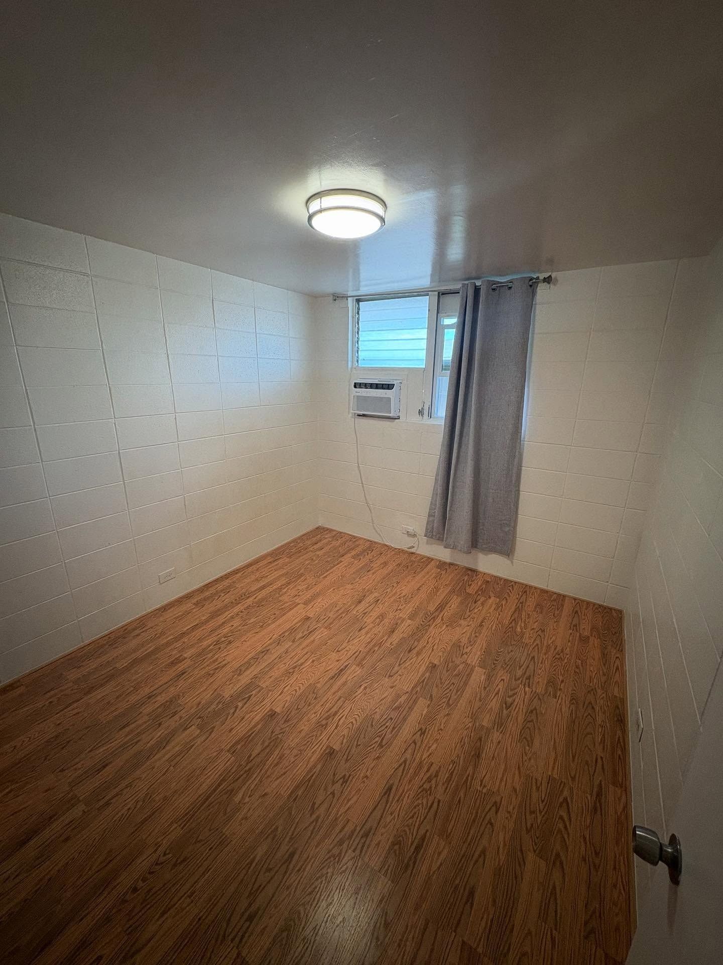 Small room with wood flooring, white walls, small window with AC unit and grey curtain, ceiling light.
