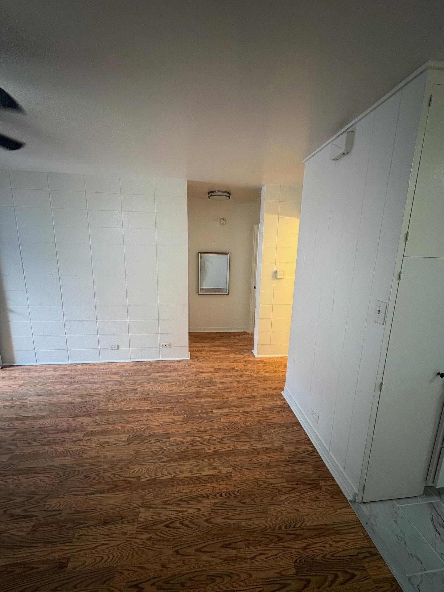 Empty room with wood-look floor, white walls, and a hallway leading to a mirror.