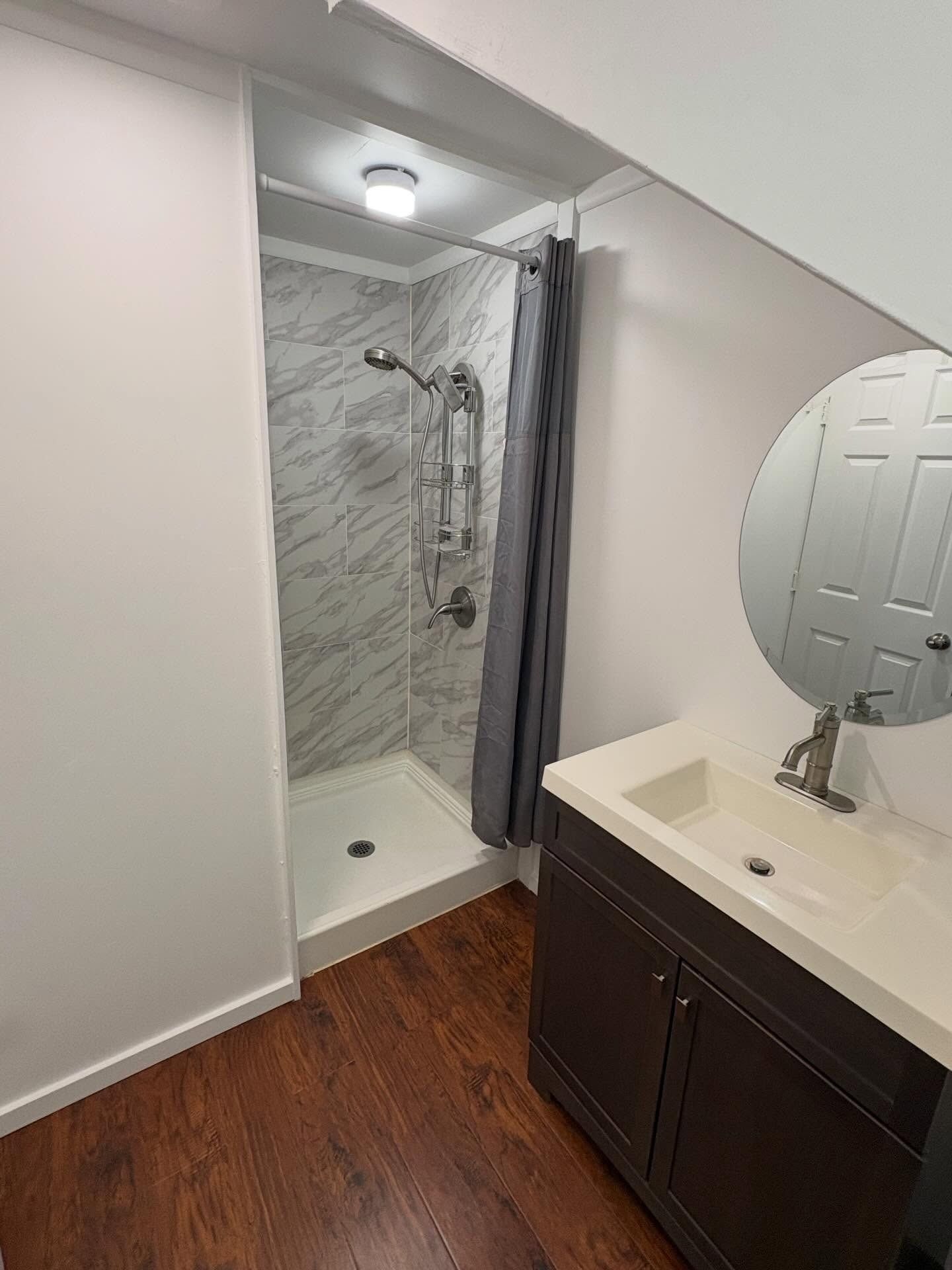 Small bathroom with shower, vanity, and round mirror. Wood-look floor, white walls, and gray shower curtain.