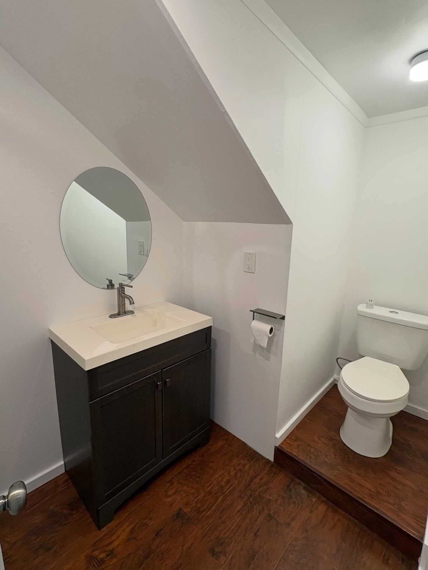 Small bathroom with vanity, round mirror, and toilet. Dark brown cabinets and flooring, white walls.