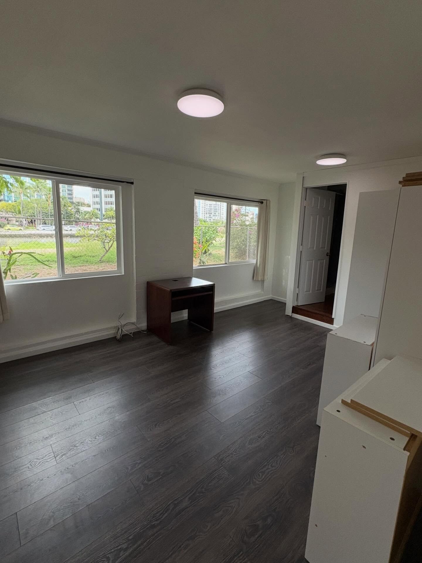 Empty room with dark wood-look flooring, two windows, desk, and white walls.