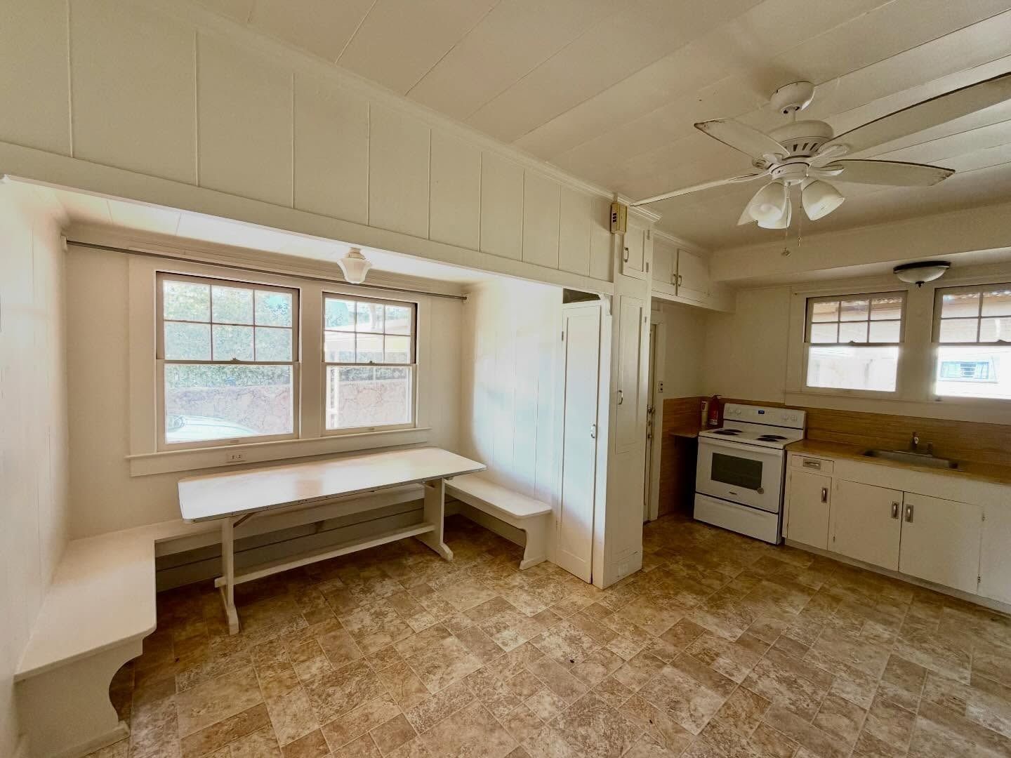 Empty kitchen with windows, stove, cabinets, and a built-in bench. White walls and ceiling, and a ceiling fan.