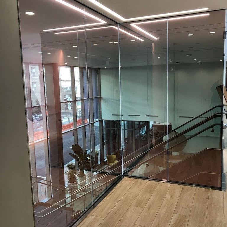 Modern indoor stairwell with glass walls, wood floor, and black metal railing in a bright building.