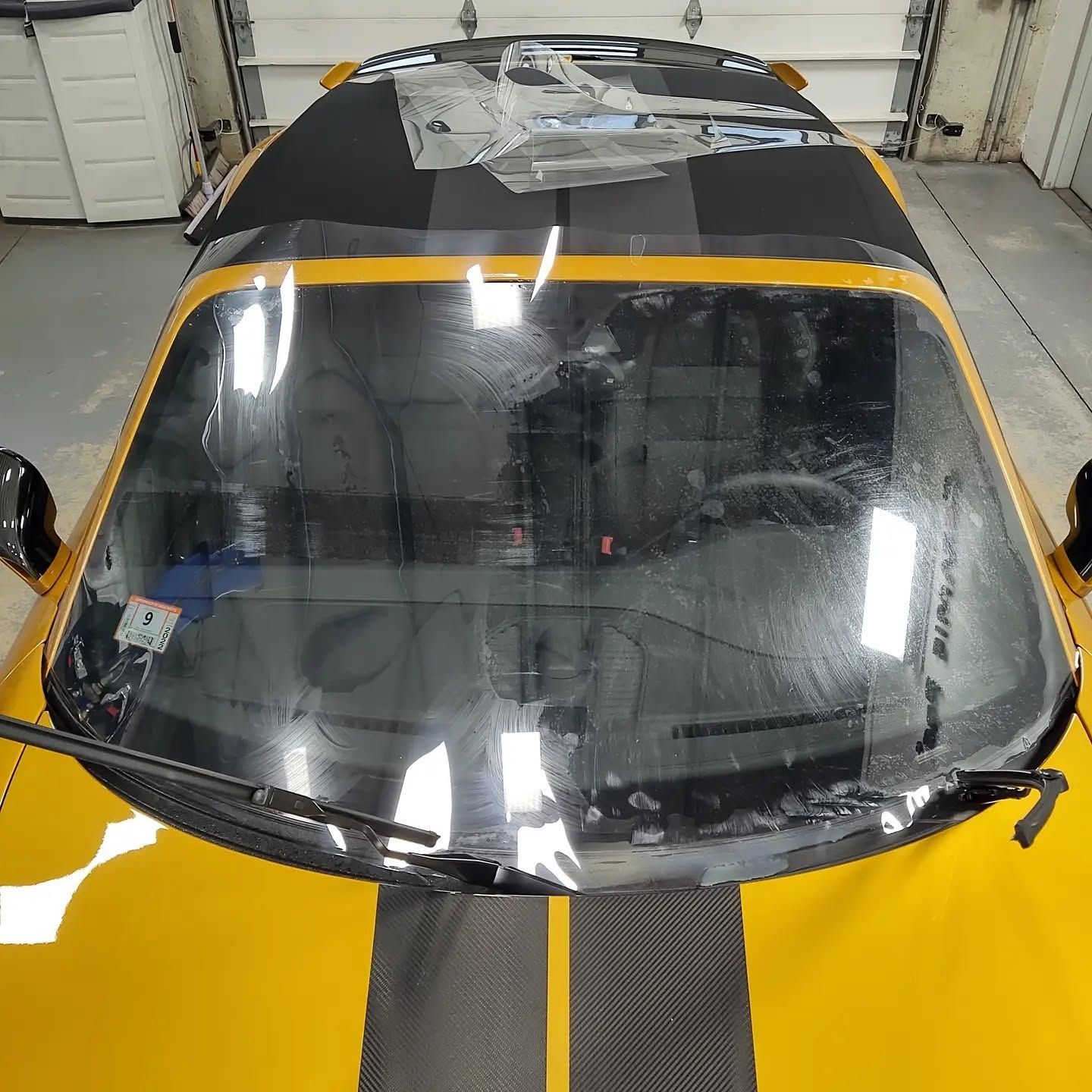 Yellow and black race car seen from above in a garage, with a taped windshield and reflective hood.