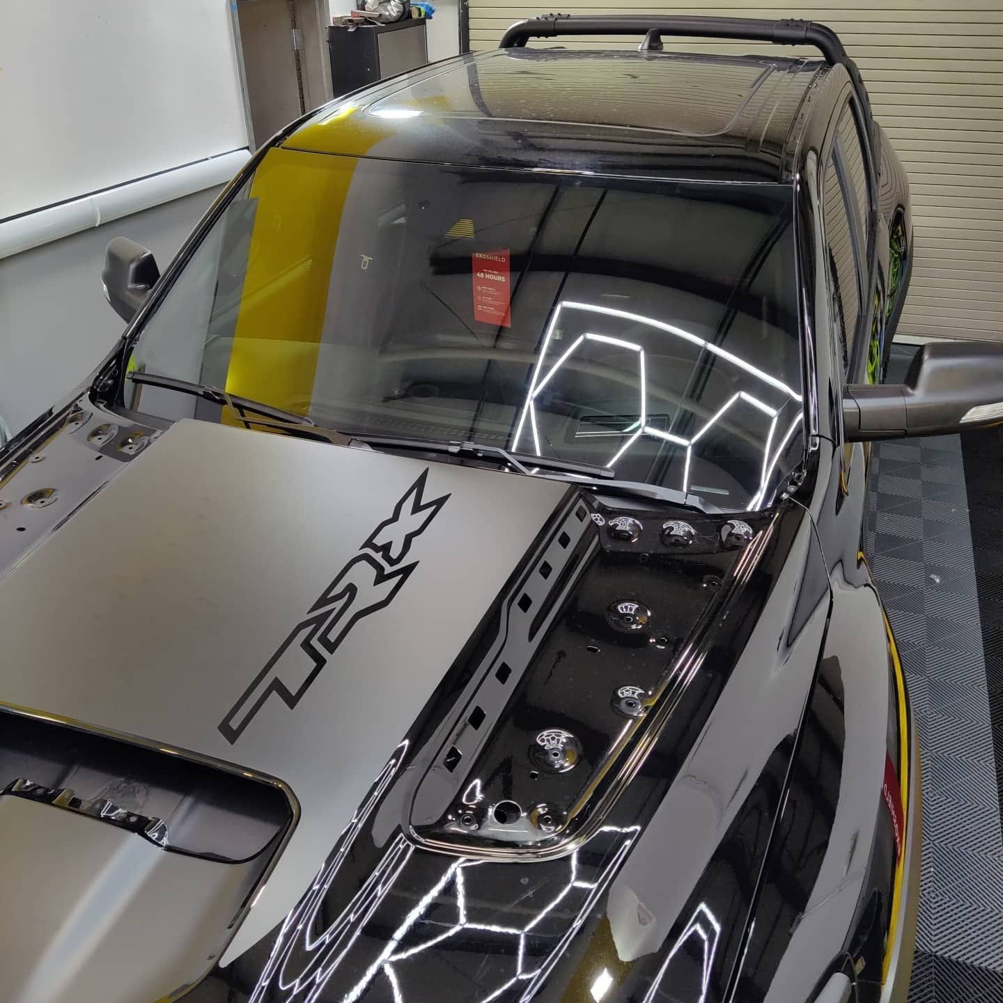 Black sports car in a garage, viewed from above, with a hood scoop and reflective windshield.