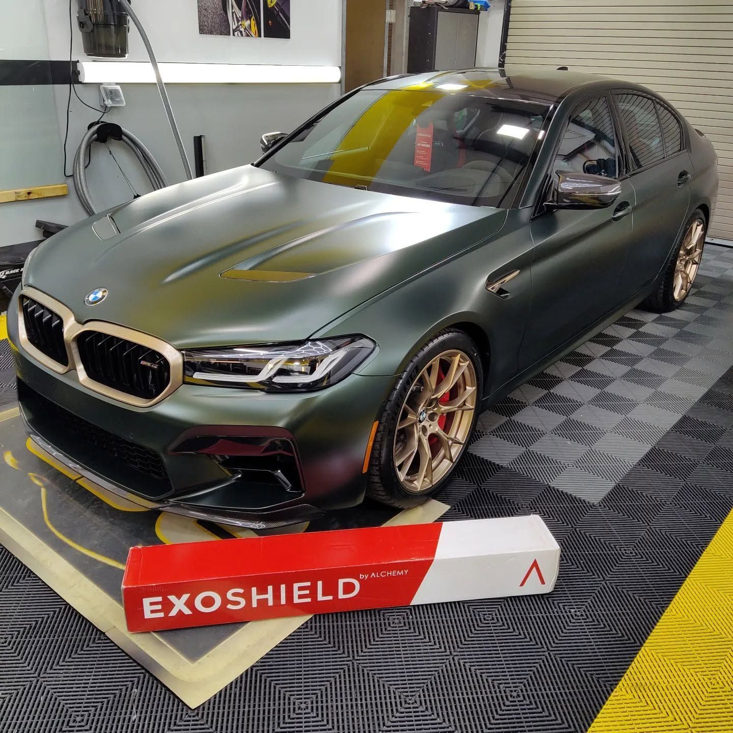 Matte green BMW coupe on display indoors, with gold wheels and an EXOSHIELD banner in front.