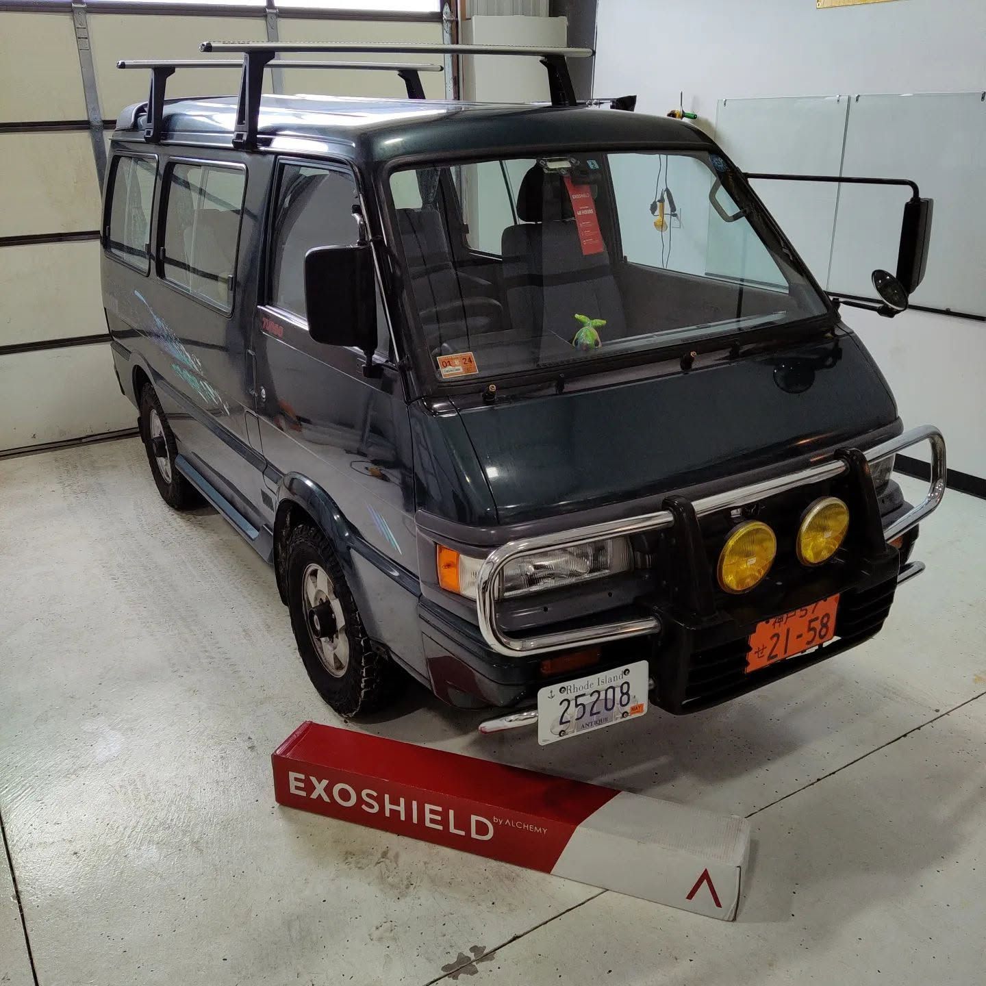 Dark green mini truck with roof rack and yellow fog lights in a garage, behind an EXOGUARD sign.