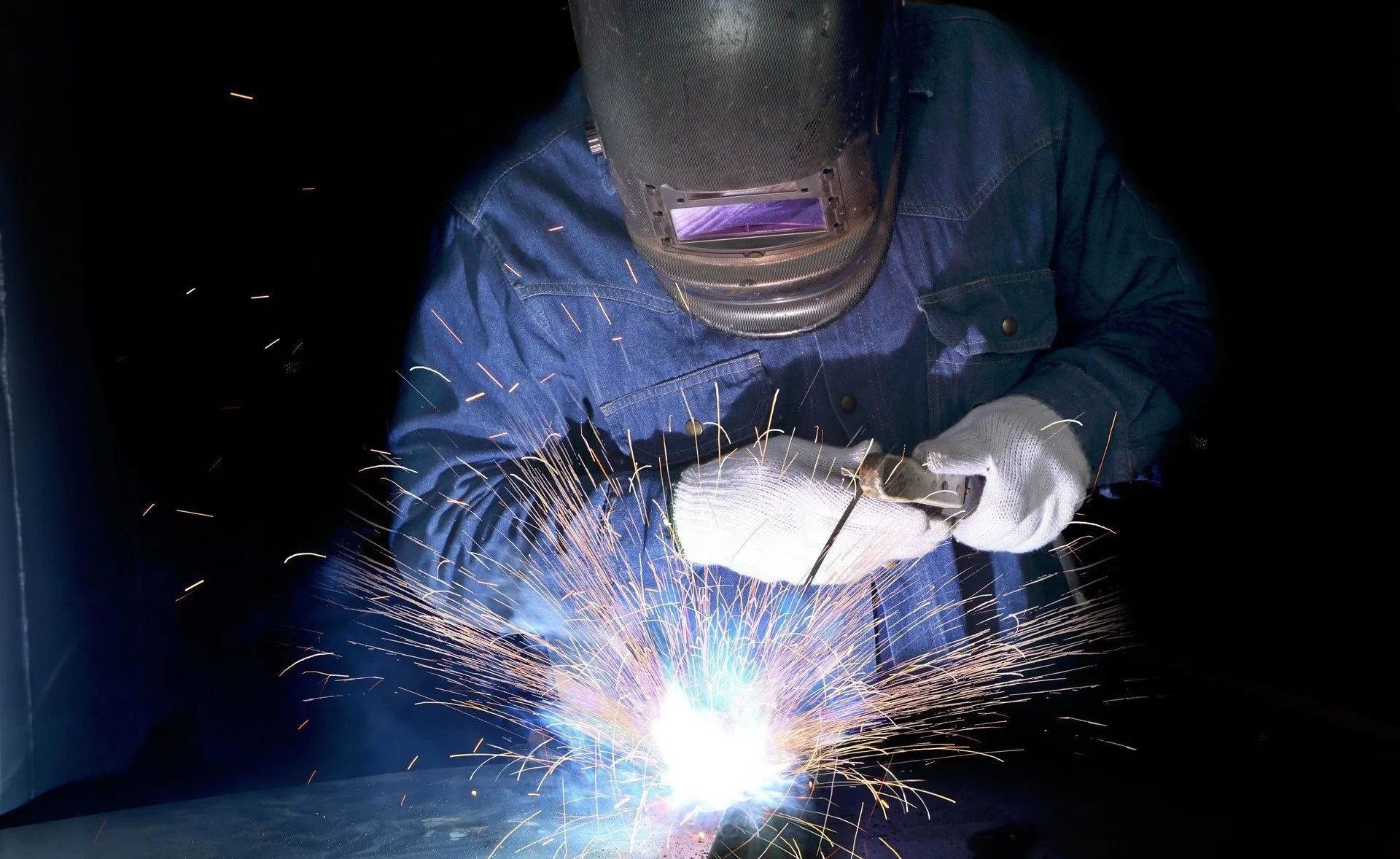 A welder in a protective mask and gloves joins metal, creating bright sparks and intense light in a dark workspace.