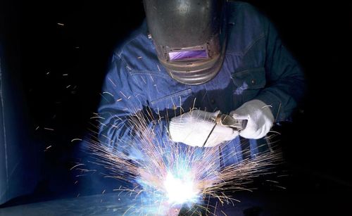 A person wearing a protective welding helmet and gloves performs metal welding, creating a bright arc and sparks.
