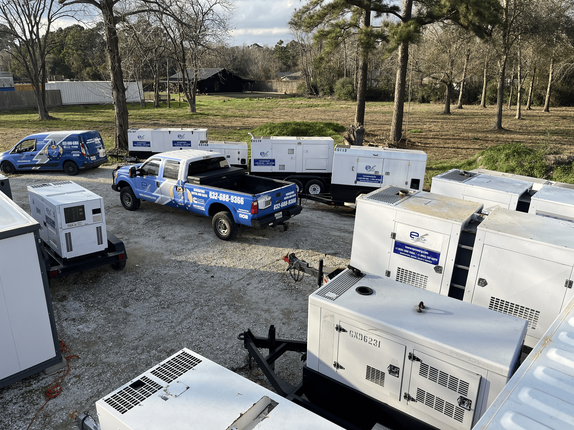 Blue trucks and white utility trailers parked on a gravel lot beside trees and grass.