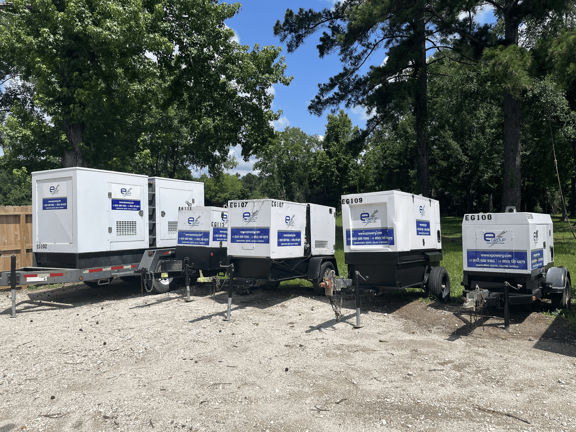 Five white portable generators on trailers lined up on gravel under trees