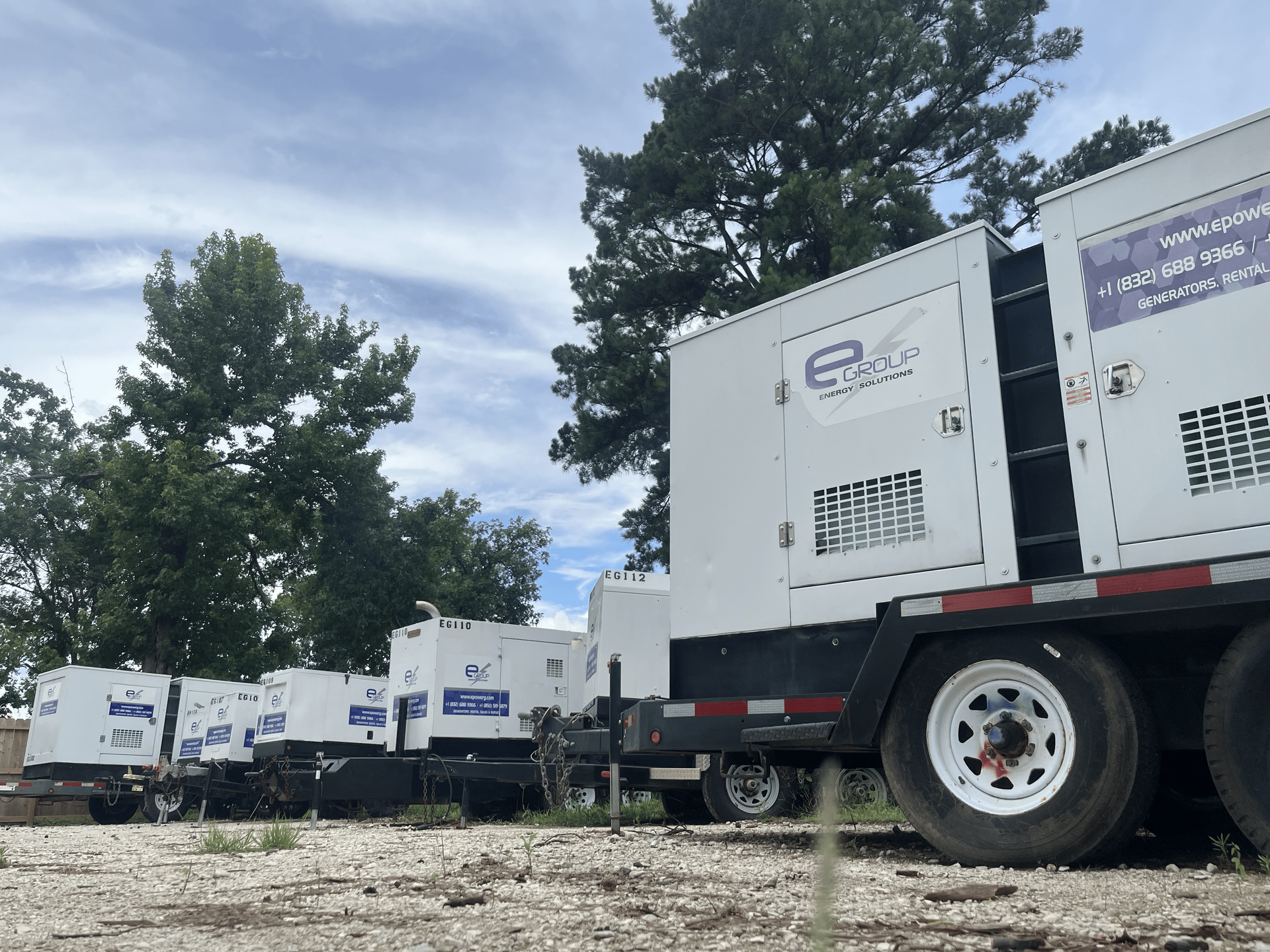 White industrial trailers with large equipment boxes parked outdoors on a gravel lot under trees and a cloudy sky
