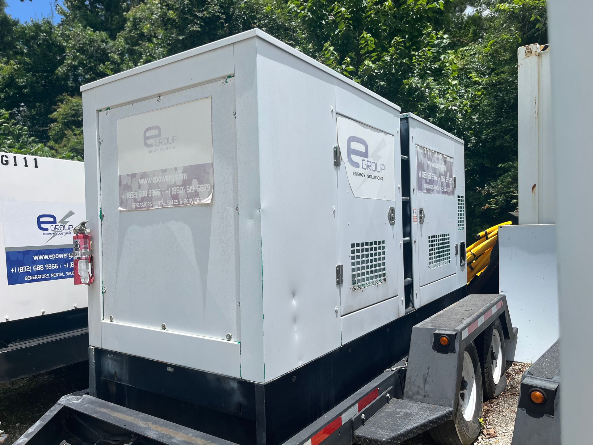 White industrial generator trailer parked outdoors beside another trailer and trees