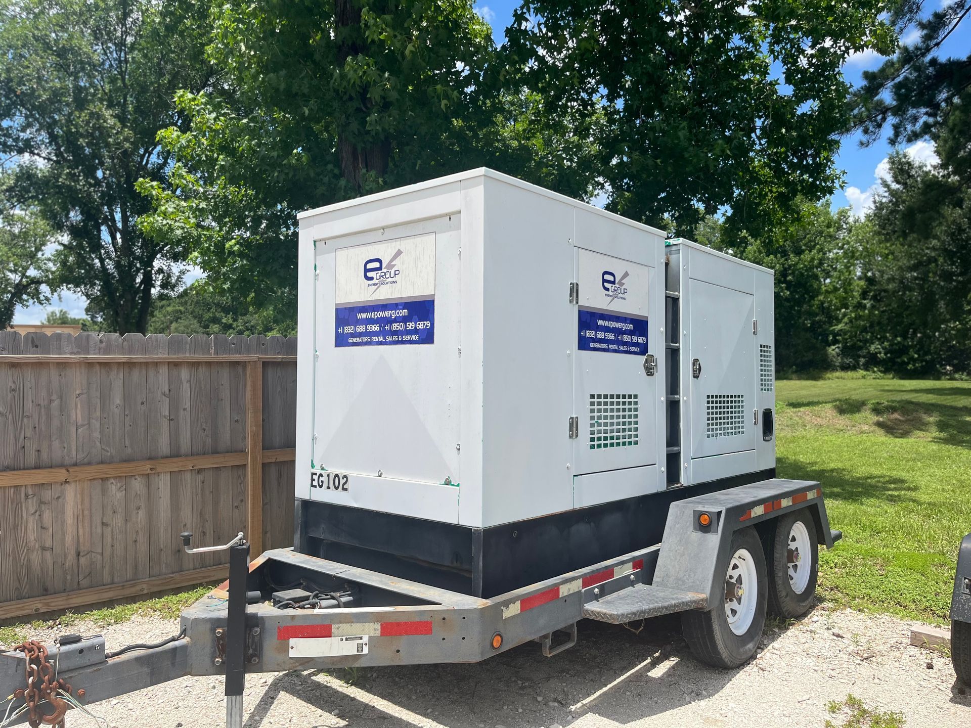 White equipment generator on a trailer parked on grass beside a fence and trees