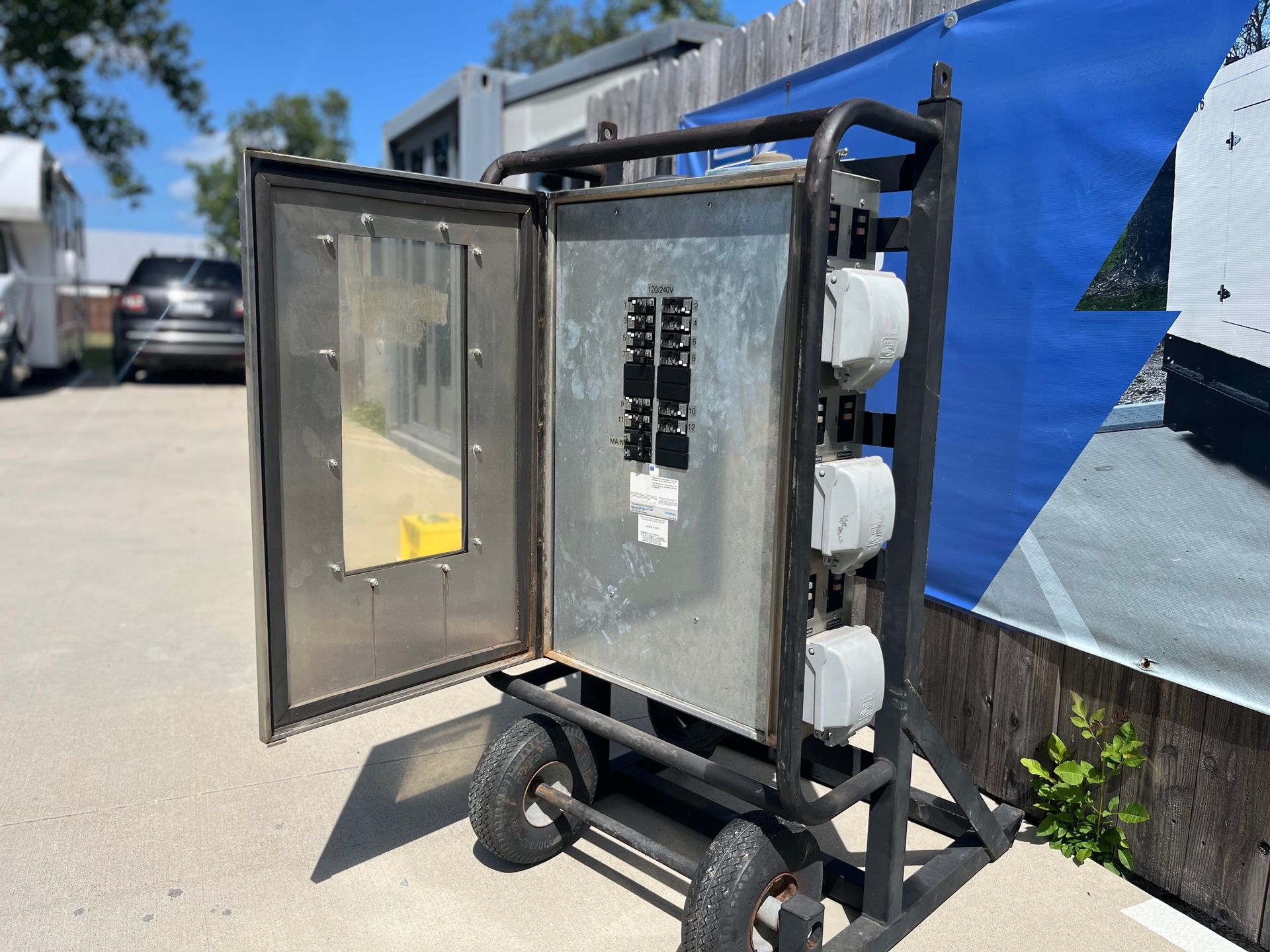 Open electrical control cabinet on a wheeled cart outdoors, with blue tarp and parked cars in the background.