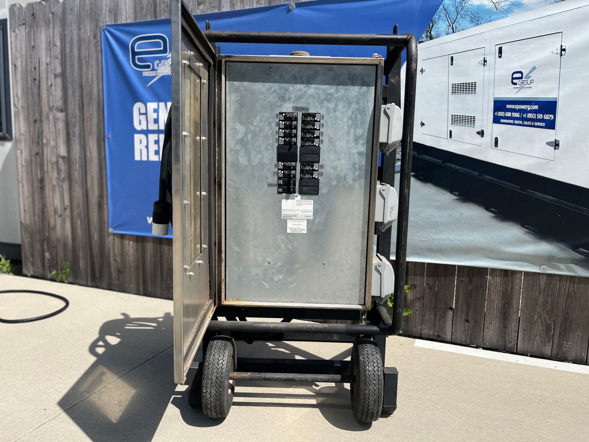 Large metal electrical cabinet on a wheeled cart beside a fence and utility truck in sunlight