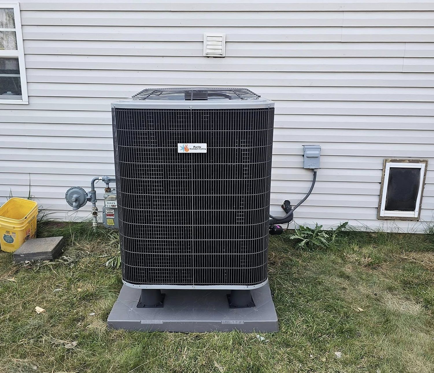 Outdoor HVAC condenser unit with black protective cover beside a house wall on a concrete pad.