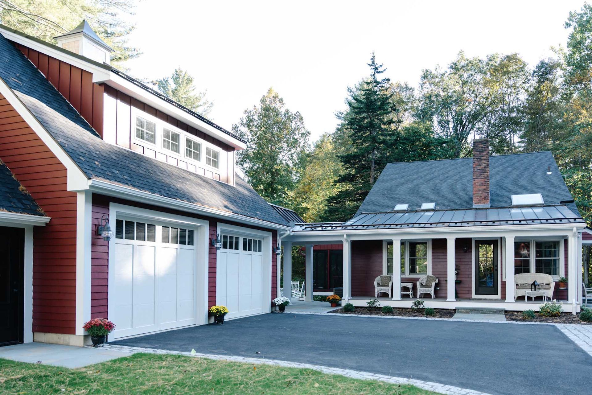 Red-sided house and garage with white doors sit on a paved driveway; porch has chairs; trees in background.
