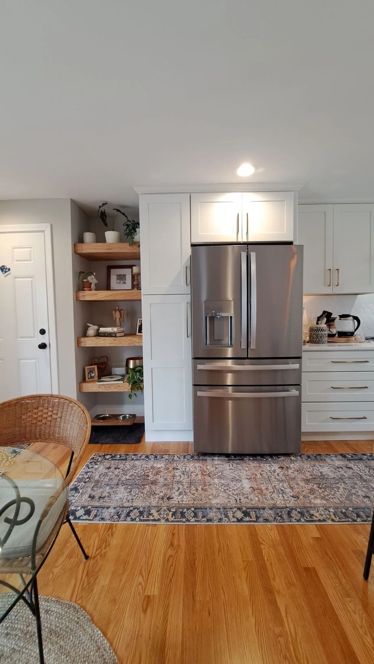 Kitchen with stainless steel refrigerator, white cabinets, open shelving, and wood flooring. A rug covers part of the floor.