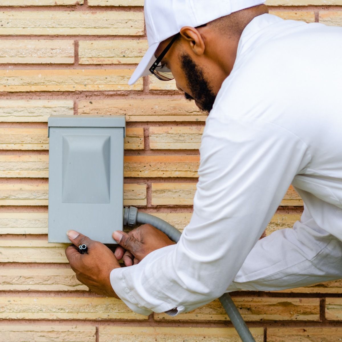 Person in white work clothes installing electrical box on brick wall.