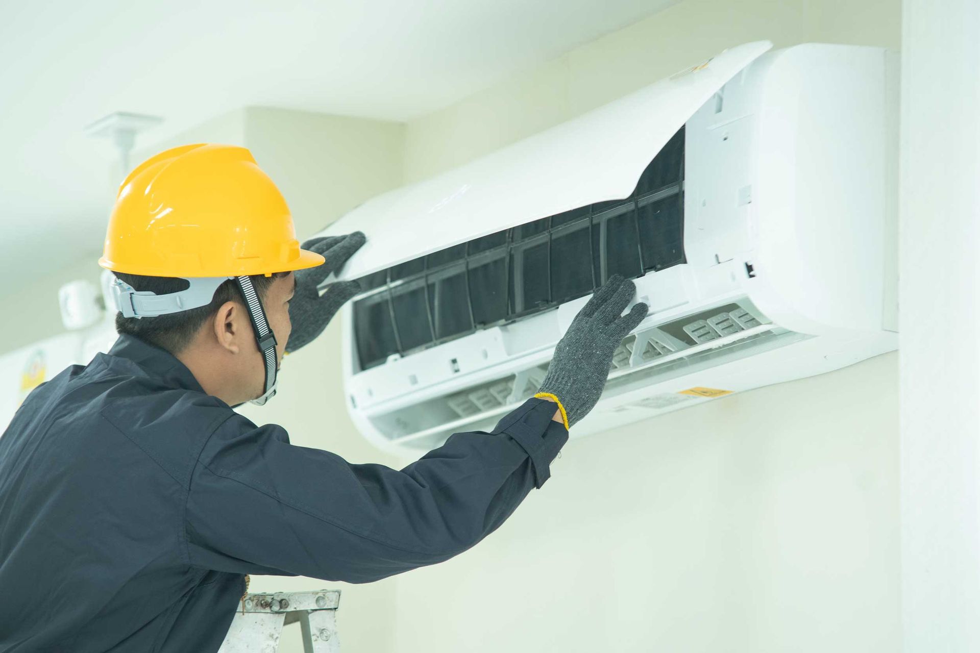 Technician in yellow hard hat cleaning a wall-mounted air conditioner indoors