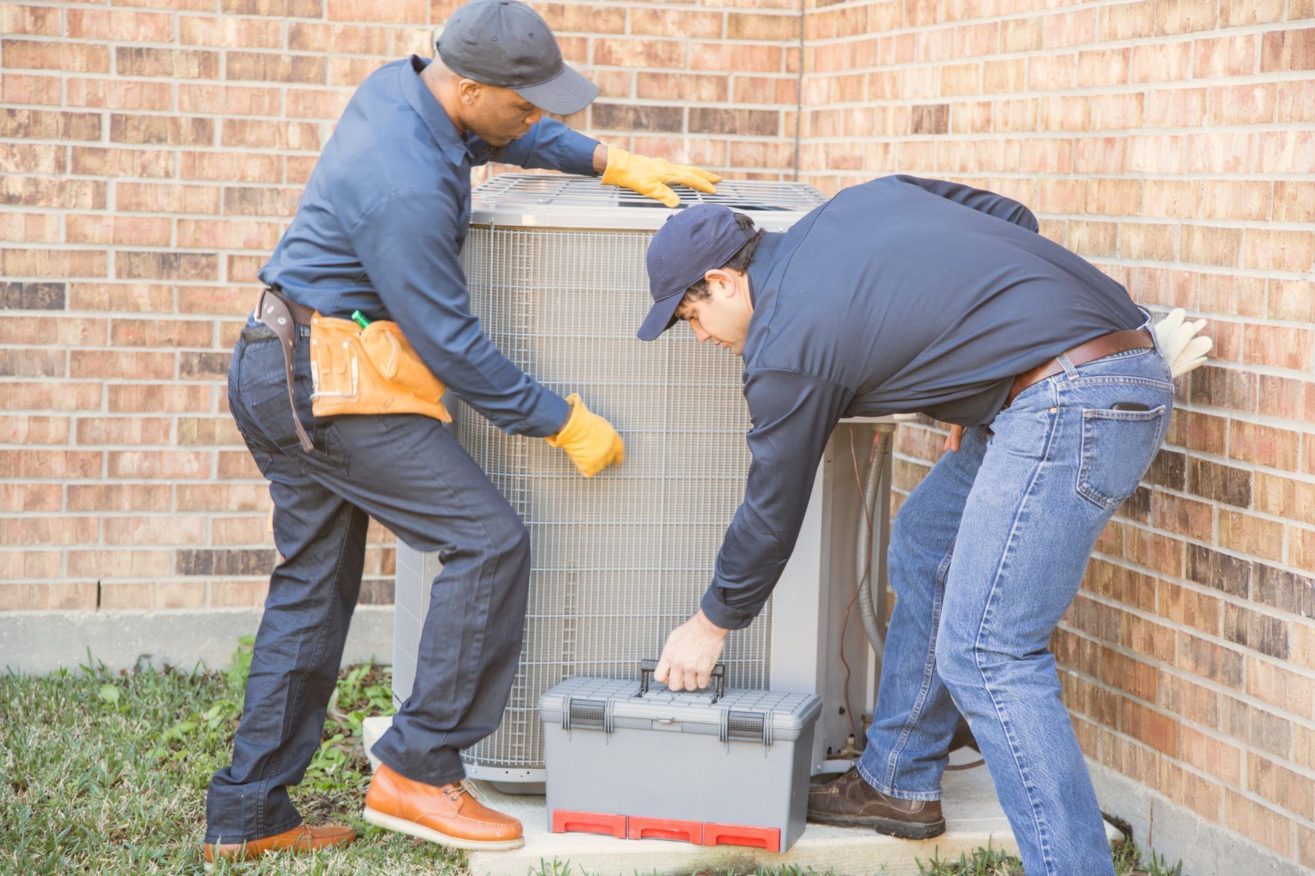Two workers carrying a toolbox beside a brick wall outdoors.
