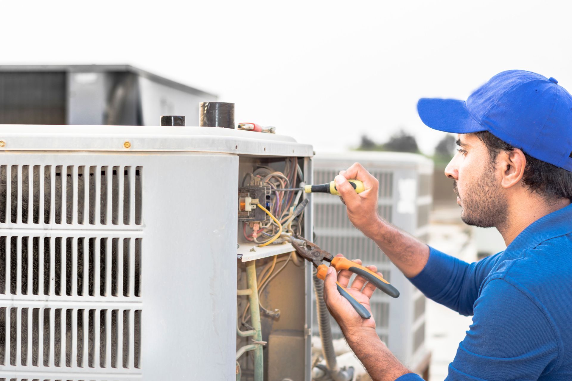 Technician in blue cap repairing exposed wiring on an outdoor HVAC unit