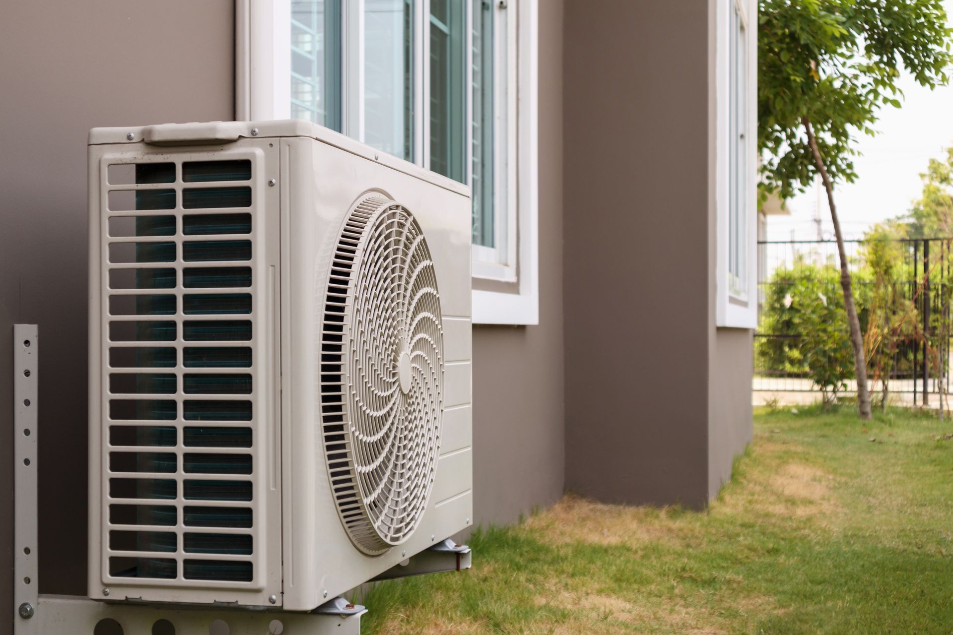 Outdoor air conditioner unit beside a house wall, with green grass and a tree in the background.
