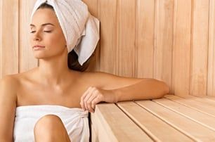 Woman wrapped in a towel, relaxing in a wooden sauna, eyes closed, leaning on a bench.