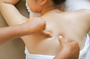Hands massaging a person's upper back, focusing on the shoulder blade area. Light skin, white towel, close-up.