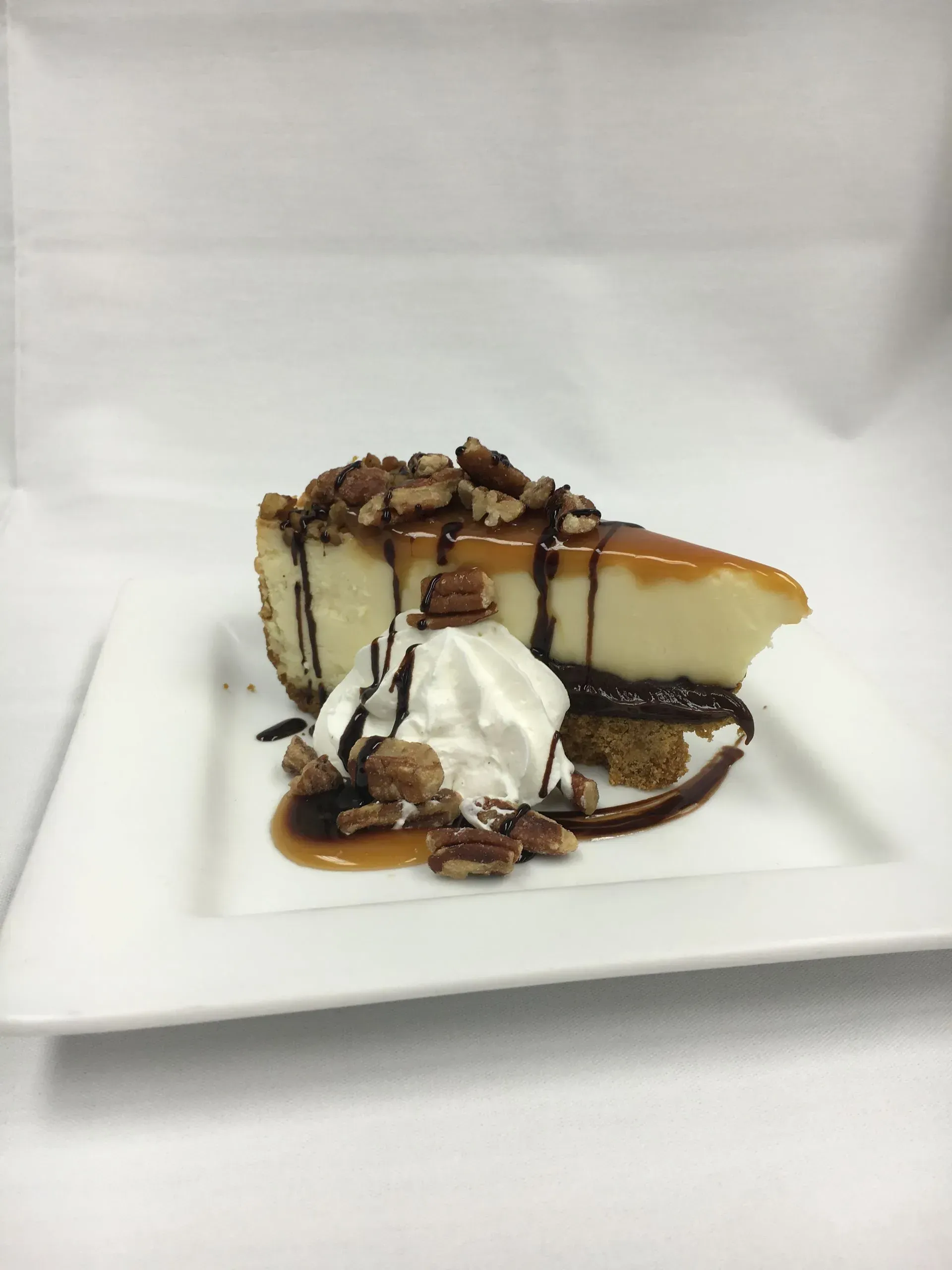 Slice of cheesecake with caramel, pecans, chocolate drizzle, and whipped cream on a white plate.