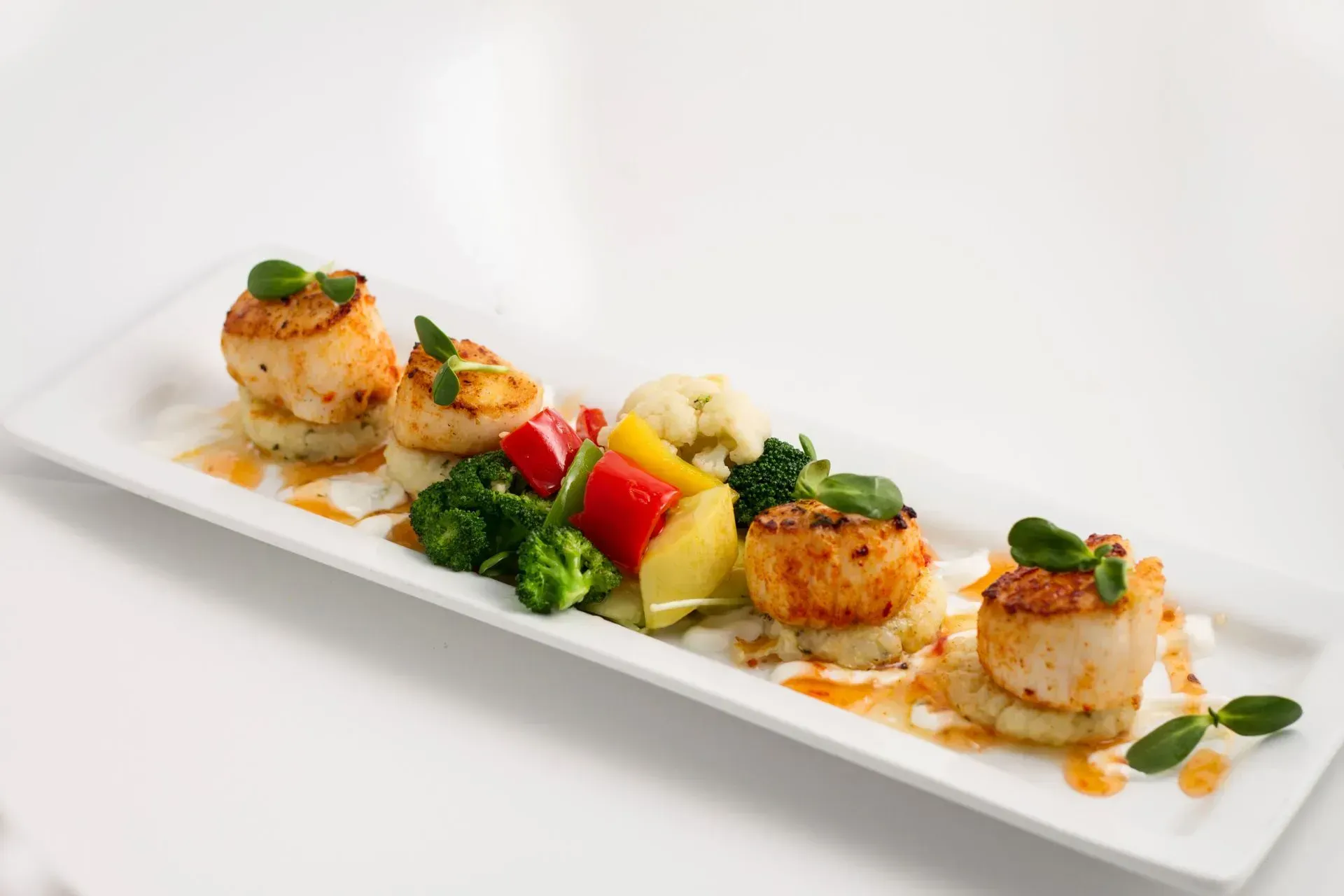 Seared scallops on mashed potatoes, with vegetables on a rectangular white plate.