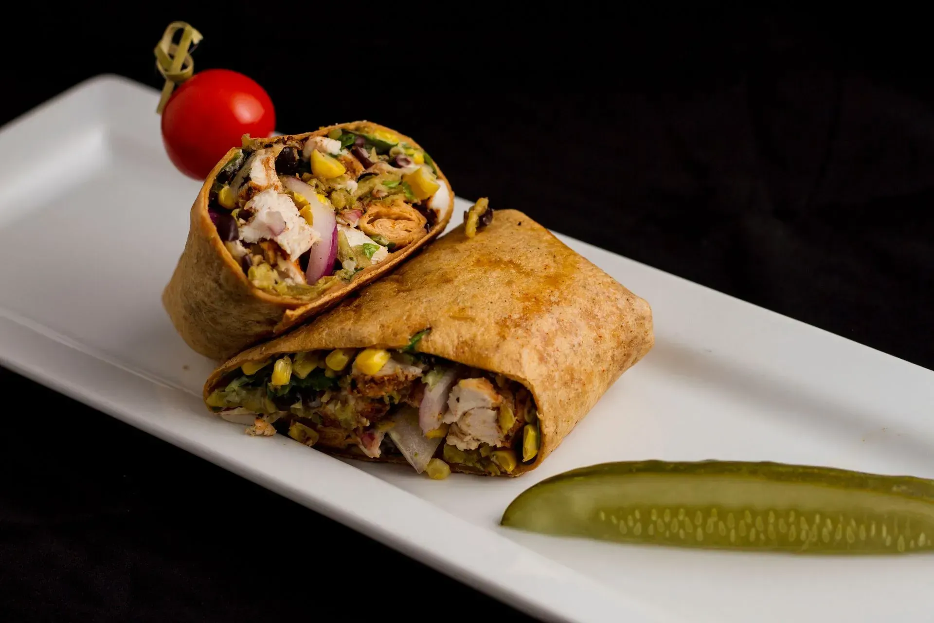 Chicken wrap on white plate with a pickle and cherry tomato garnish.