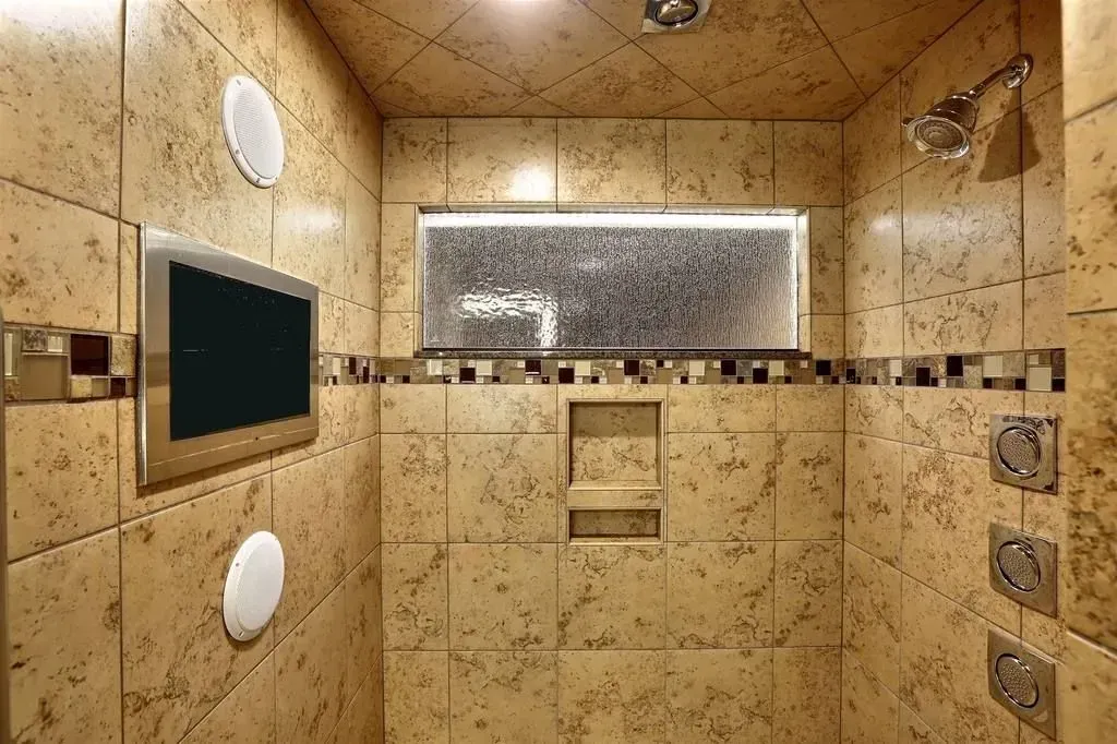 Bathroom with tiled walls, shower head, recessed shelf, and a television.