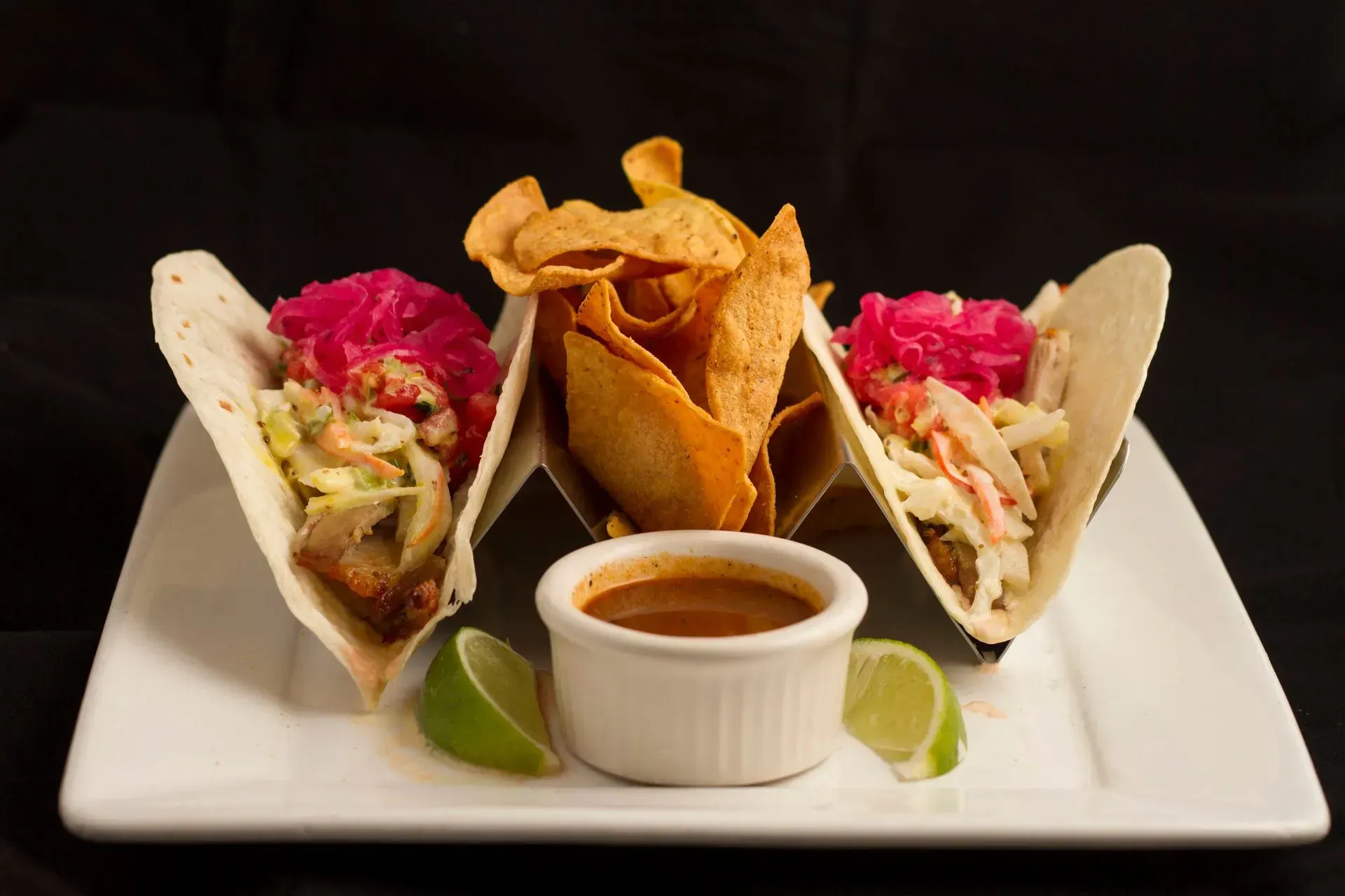 Tacos with chips and dipping sauce on a white plate. Pink pickled onions top the tacos.