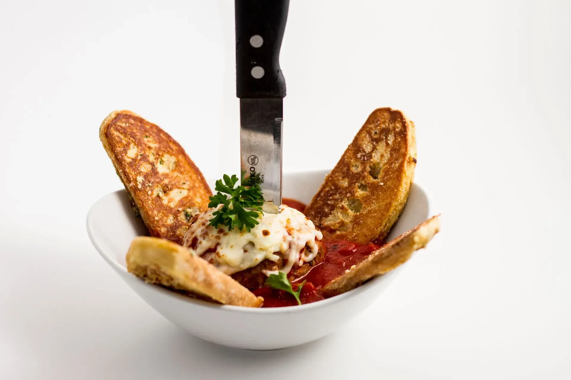 Meatball in red sauce, topped with cheese, inside a white bowl. Surrounded by toasted bread, with knife stabbed into the meatball.