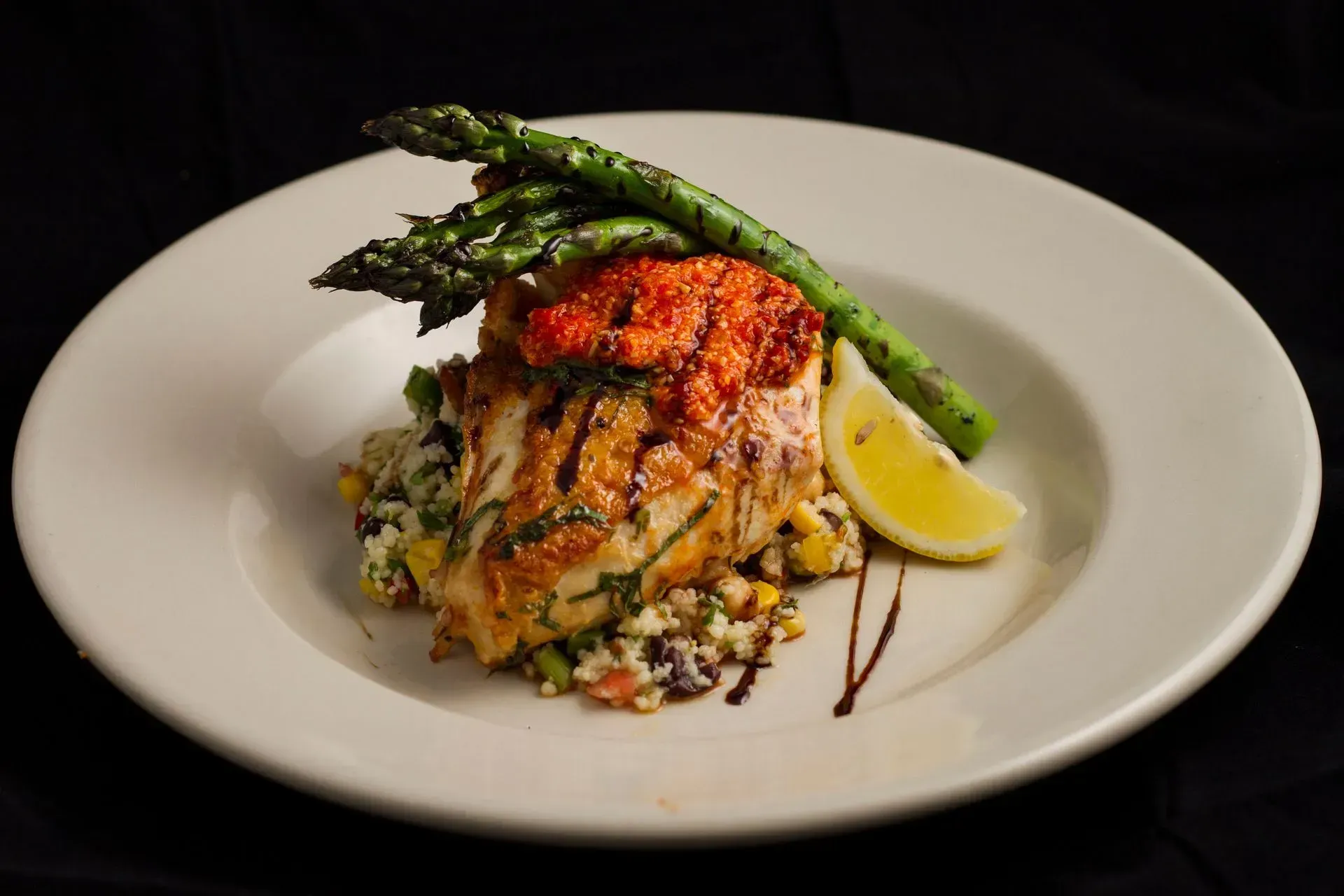 Grilled fish topped with red sauce, asparagus, and lemon on a bed of grain with balsamic drizzle.