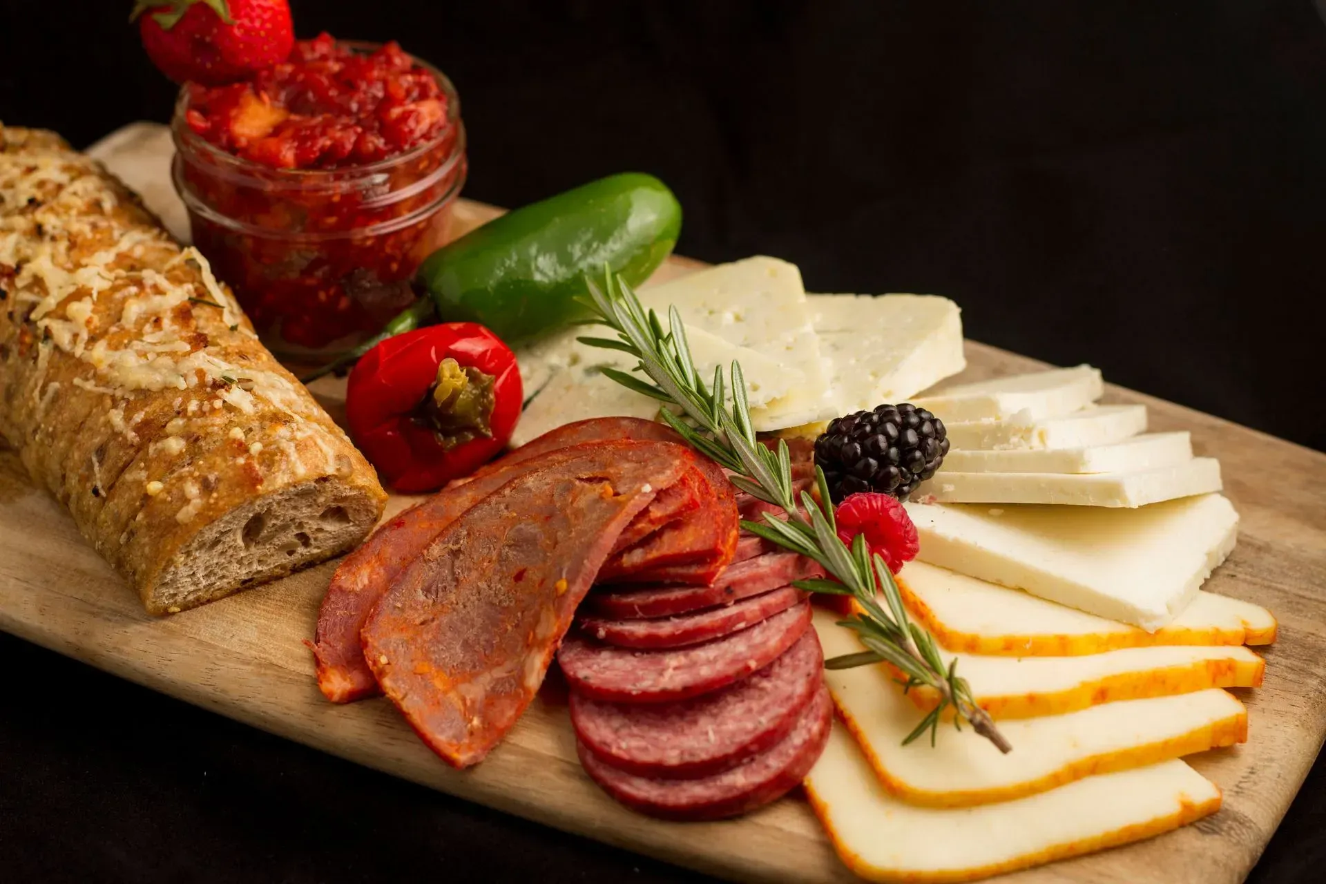 Charcuterie board with bread, salami, cheese, pepper jelly, peppers, and rosemary sprig.