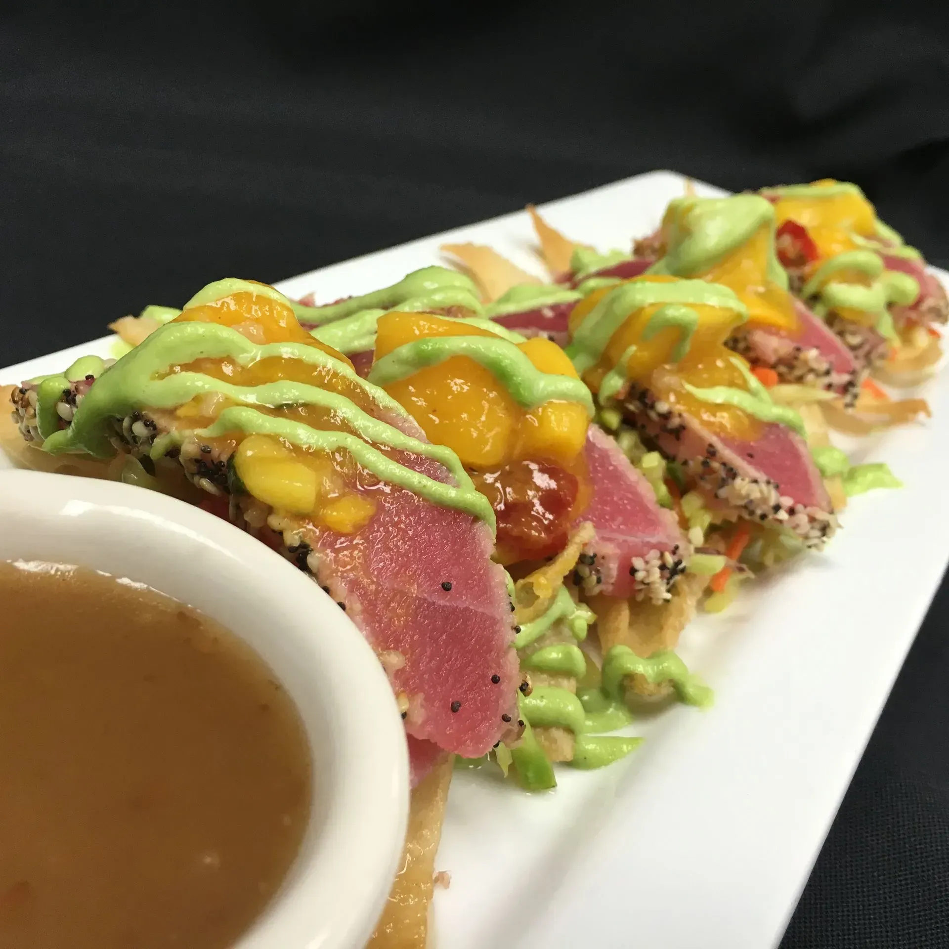 Seared tuna tacos with mango salsa and avocado crema on a white plate, served with dipping sauce.