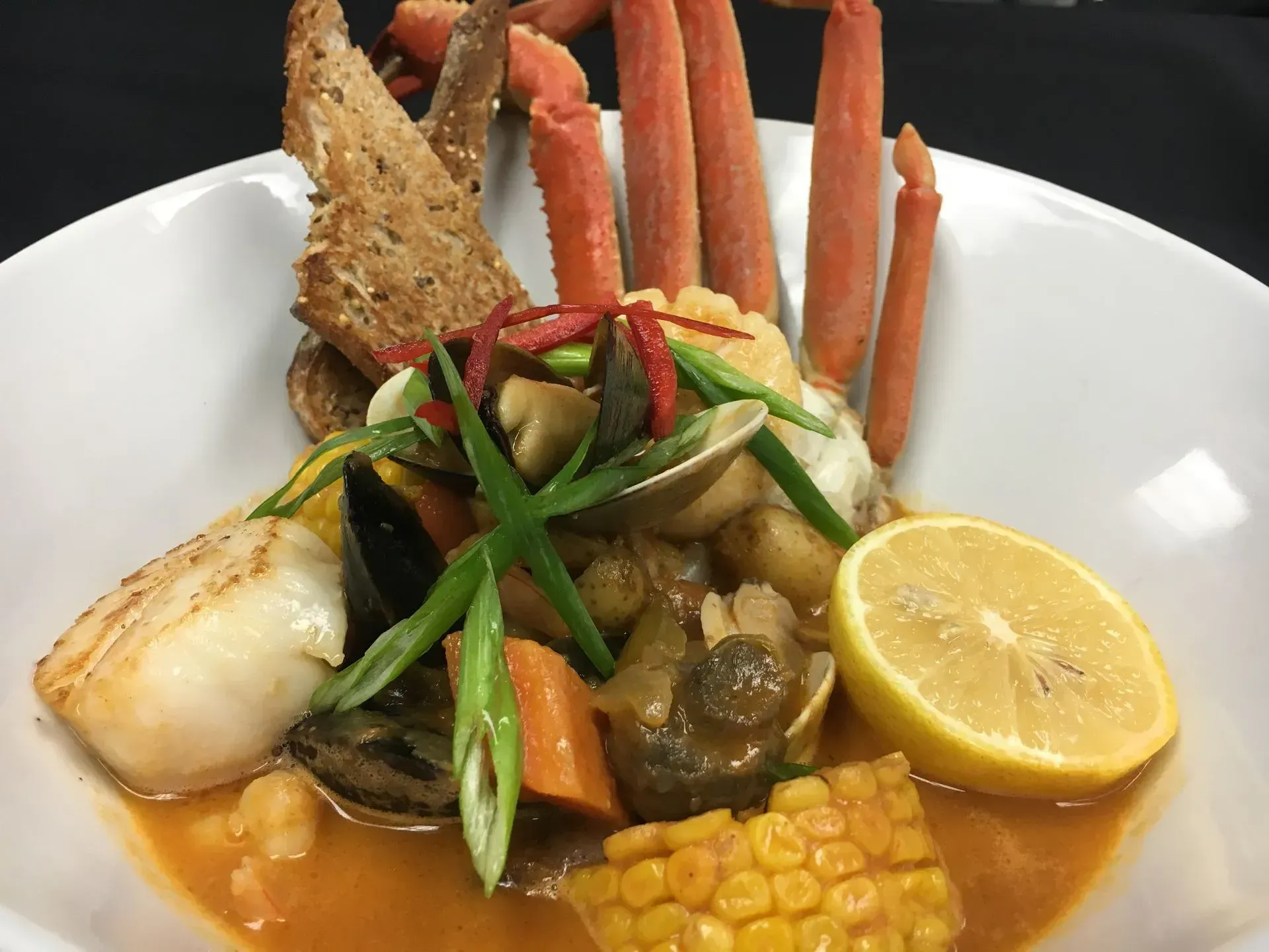 Seafood stew in a white bowl, with crab legs, scallop, vegetables, bread and a lemon wedge.