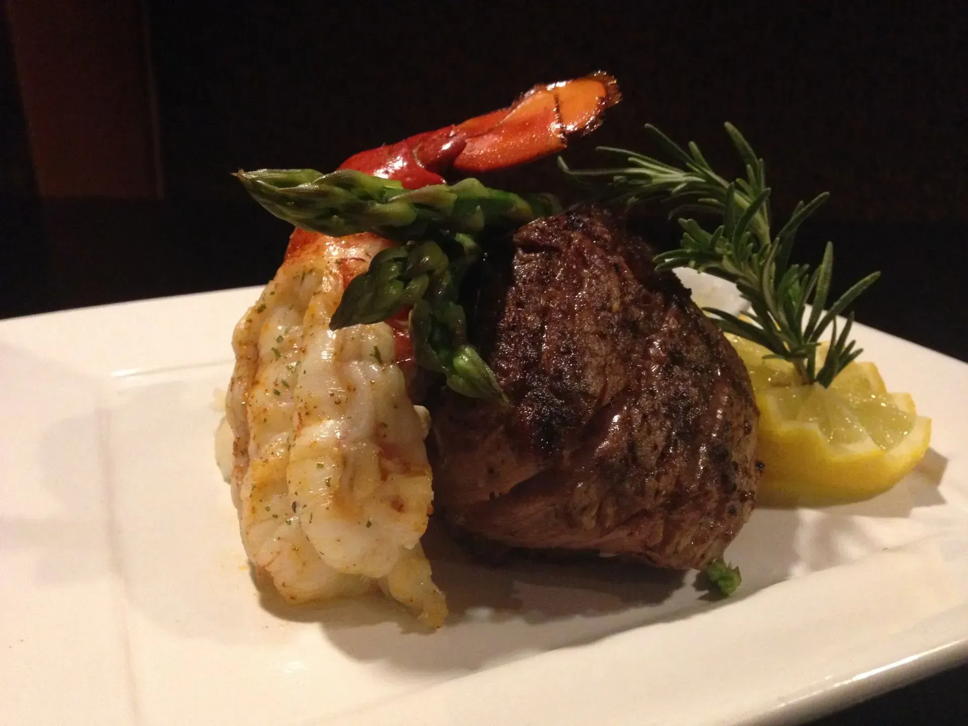 Surf and turf: grilled steak, lobster tail, asparagus, lemon, and rosemary on a white plate.