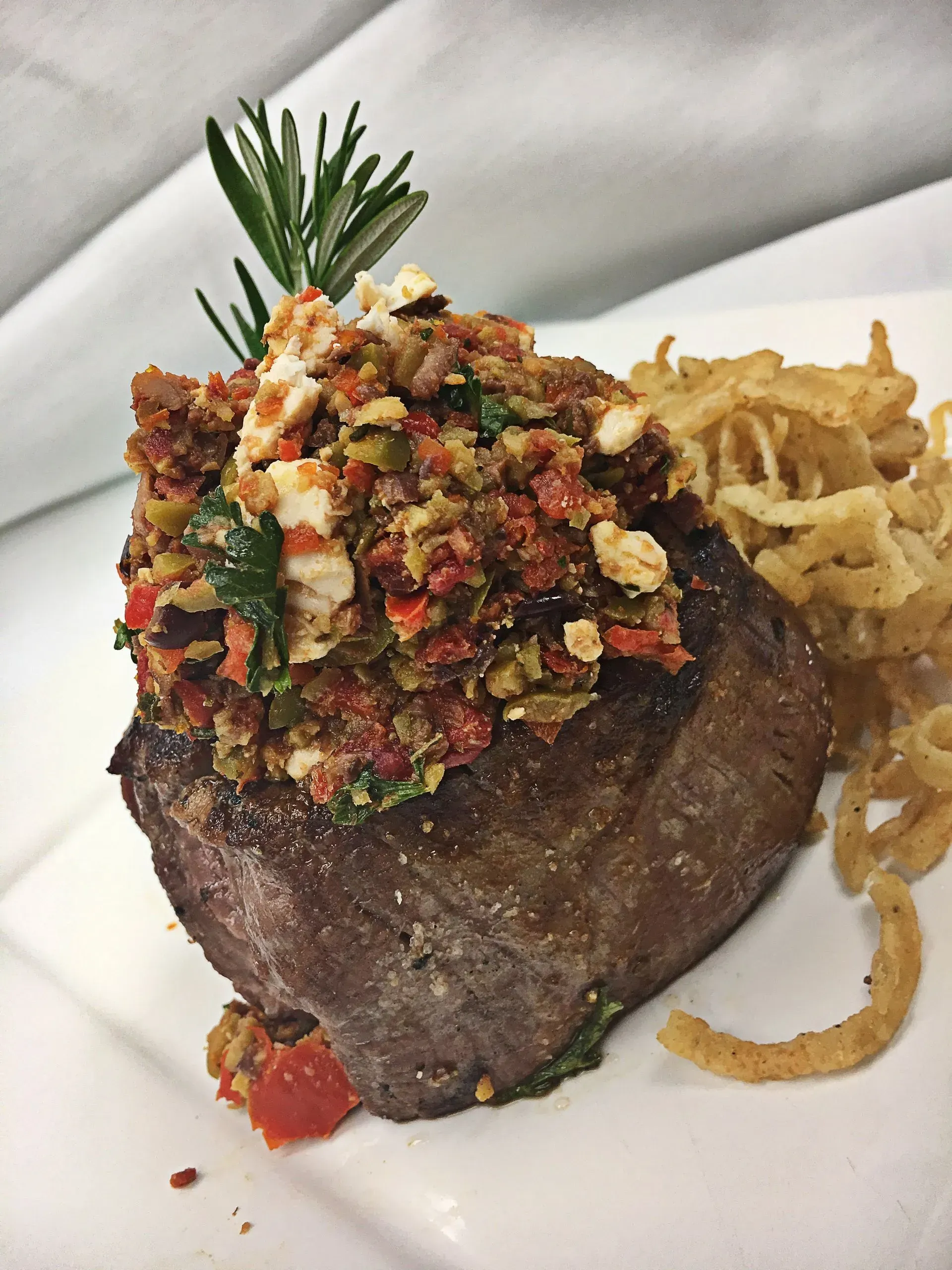 Filet mignon with a topping, served with onion rings and rosemary sprig garnish.