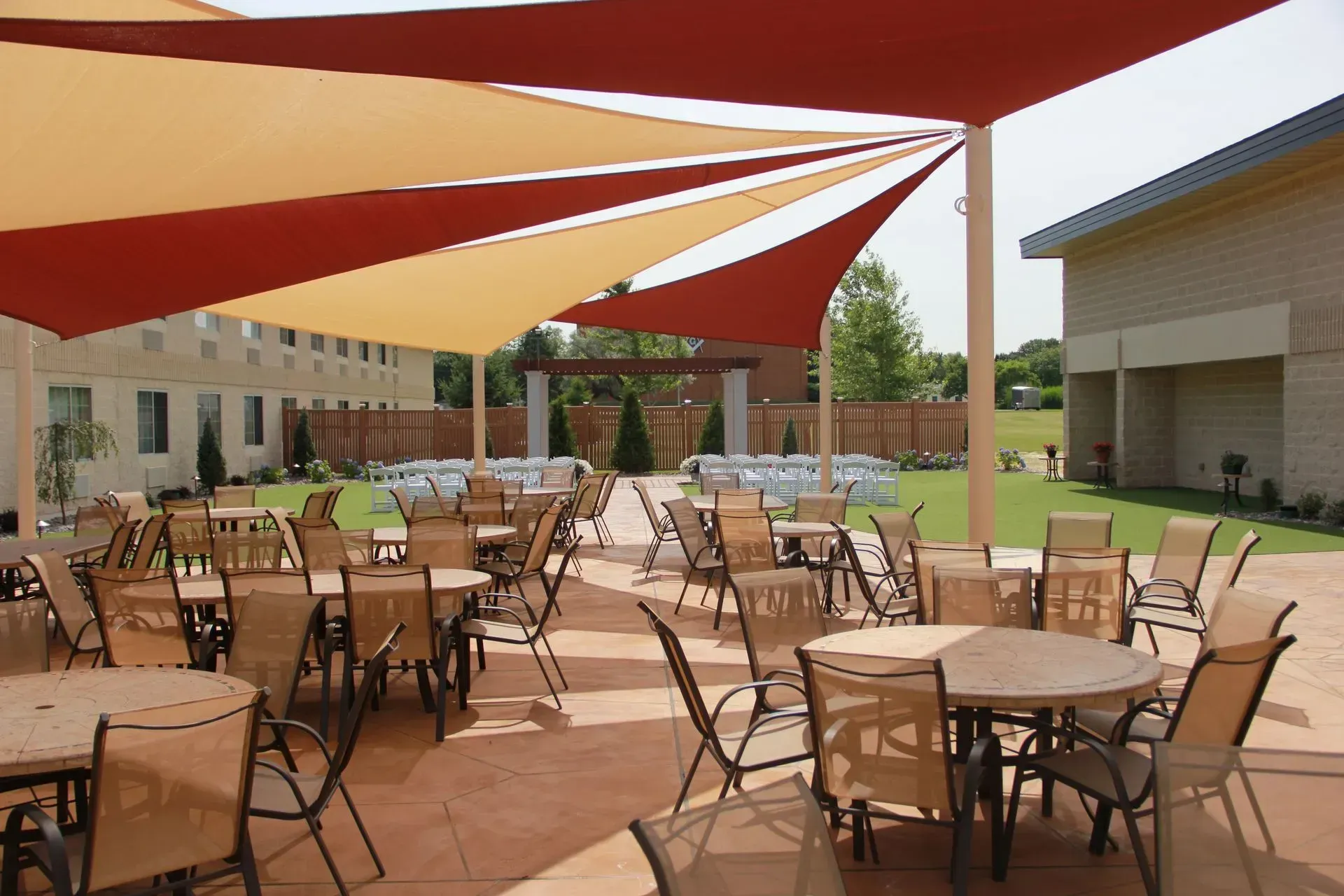 Outdoor patio with tables and chairs under a red and beige shade sail, with a building in the background.