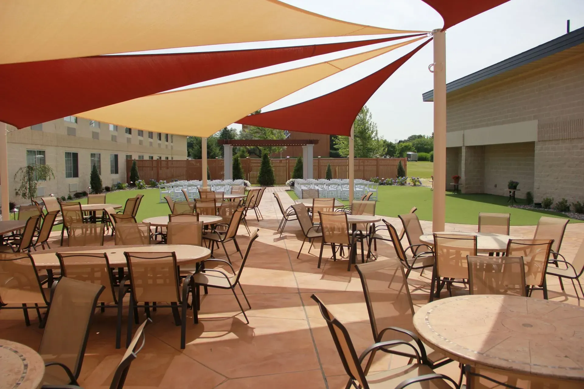Outdoor patio with tables, chairs, and shade sails; lawn, building in background.