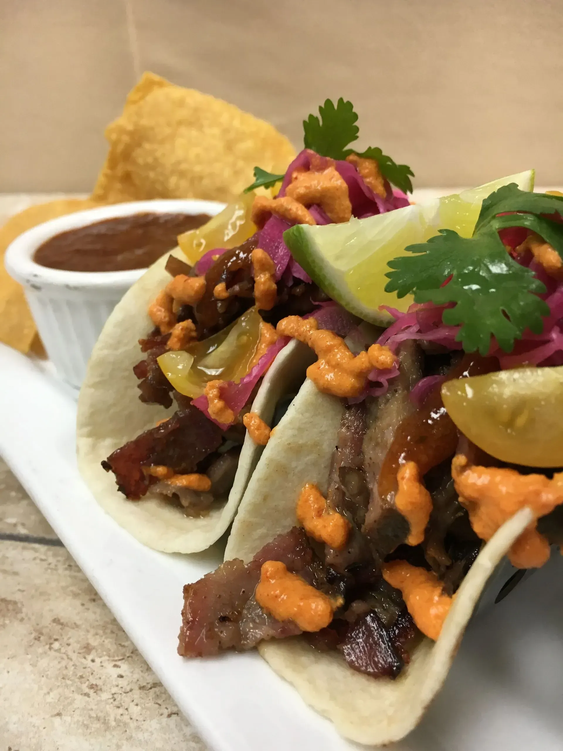 Two tacos with meat, topped with sauce, lime, and cilantro, served with salsa and chips.